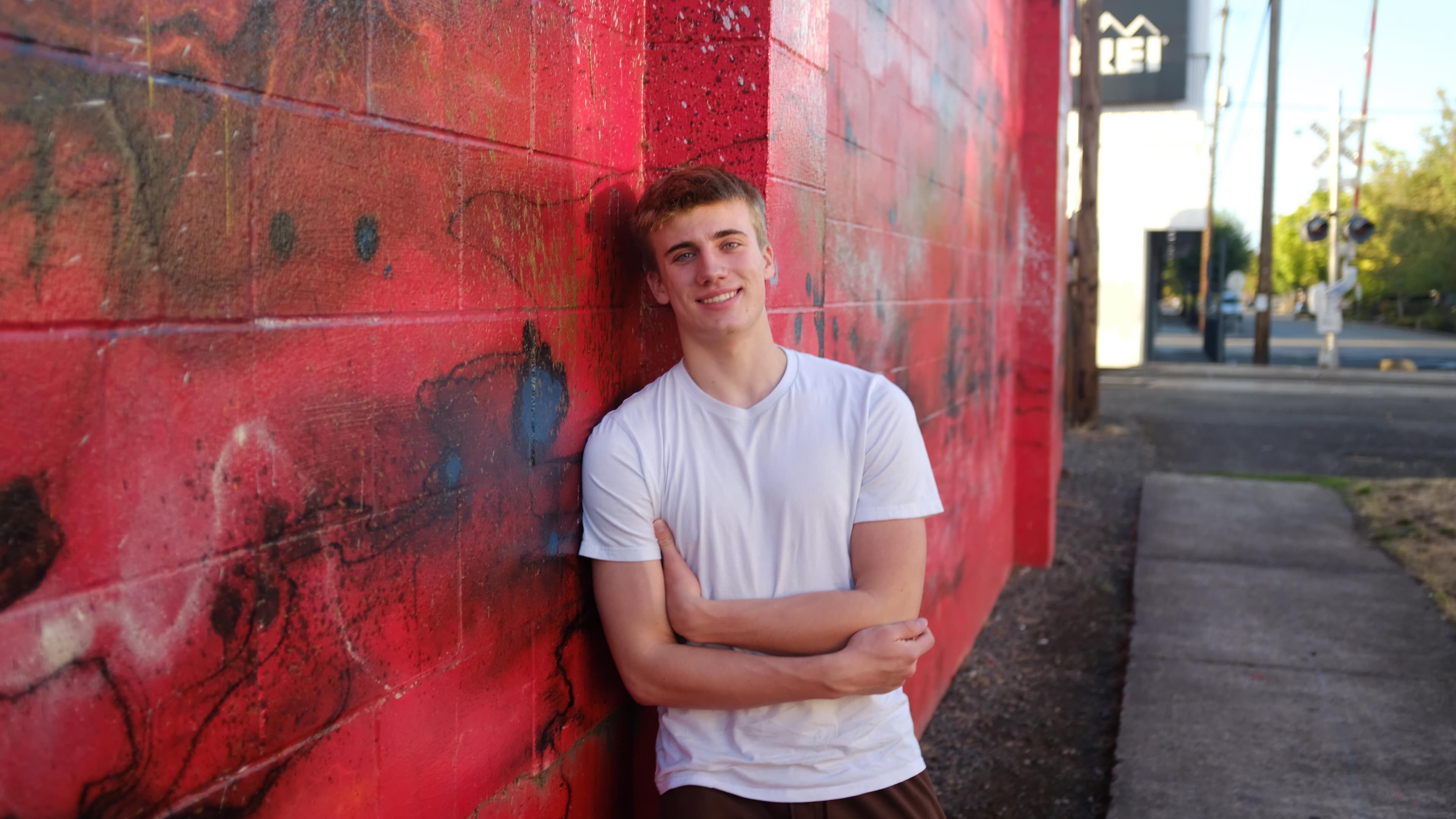 AJ Stefely, leaning against a red painted brick wall in a white t-shirt.